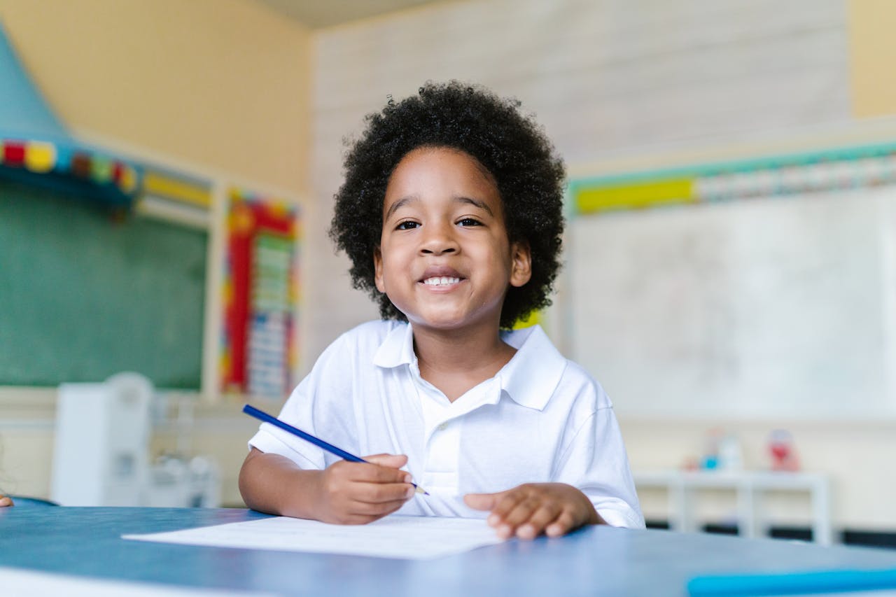 heros-img Happy child with afro hair sitting in a classroom, holding a pencil and smiling.
